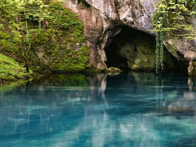 Agua serena y azul de manantial que refleja la entrada a una cueva rocosa